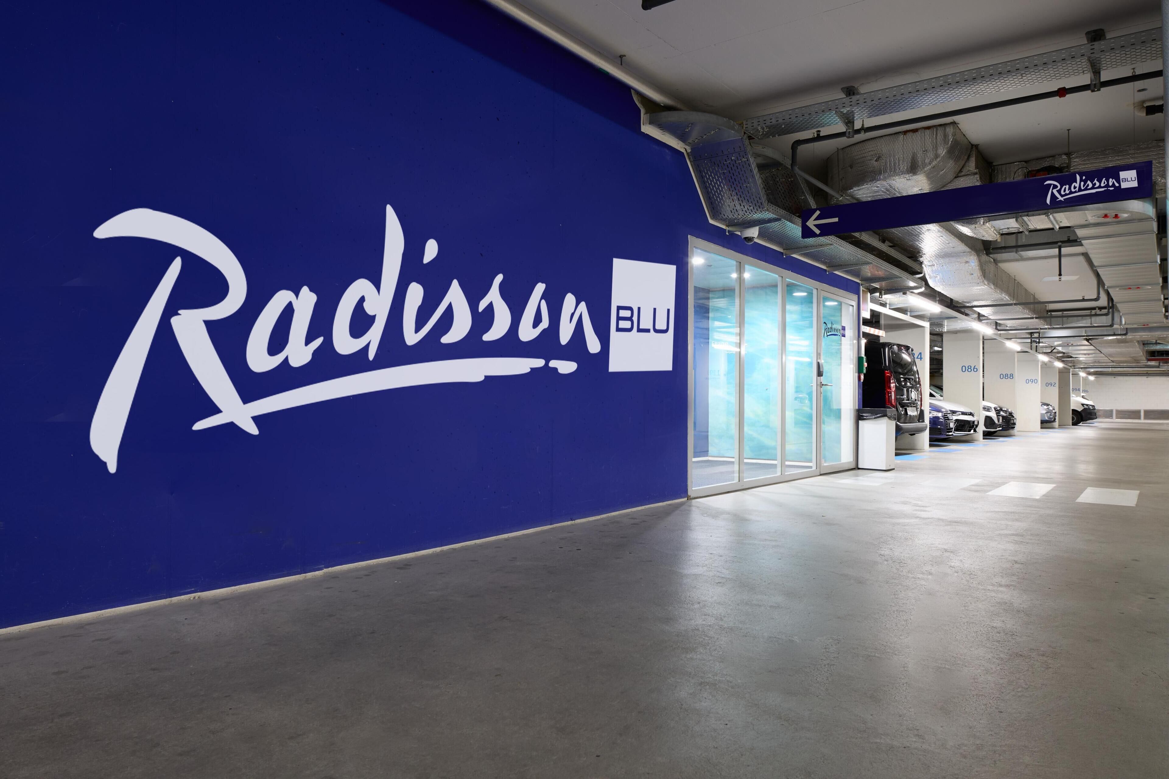 A Radisson BLU hotel corridor with a blue wall, a sign indicating the direction to Radisson BLU, and a parking area with cars.