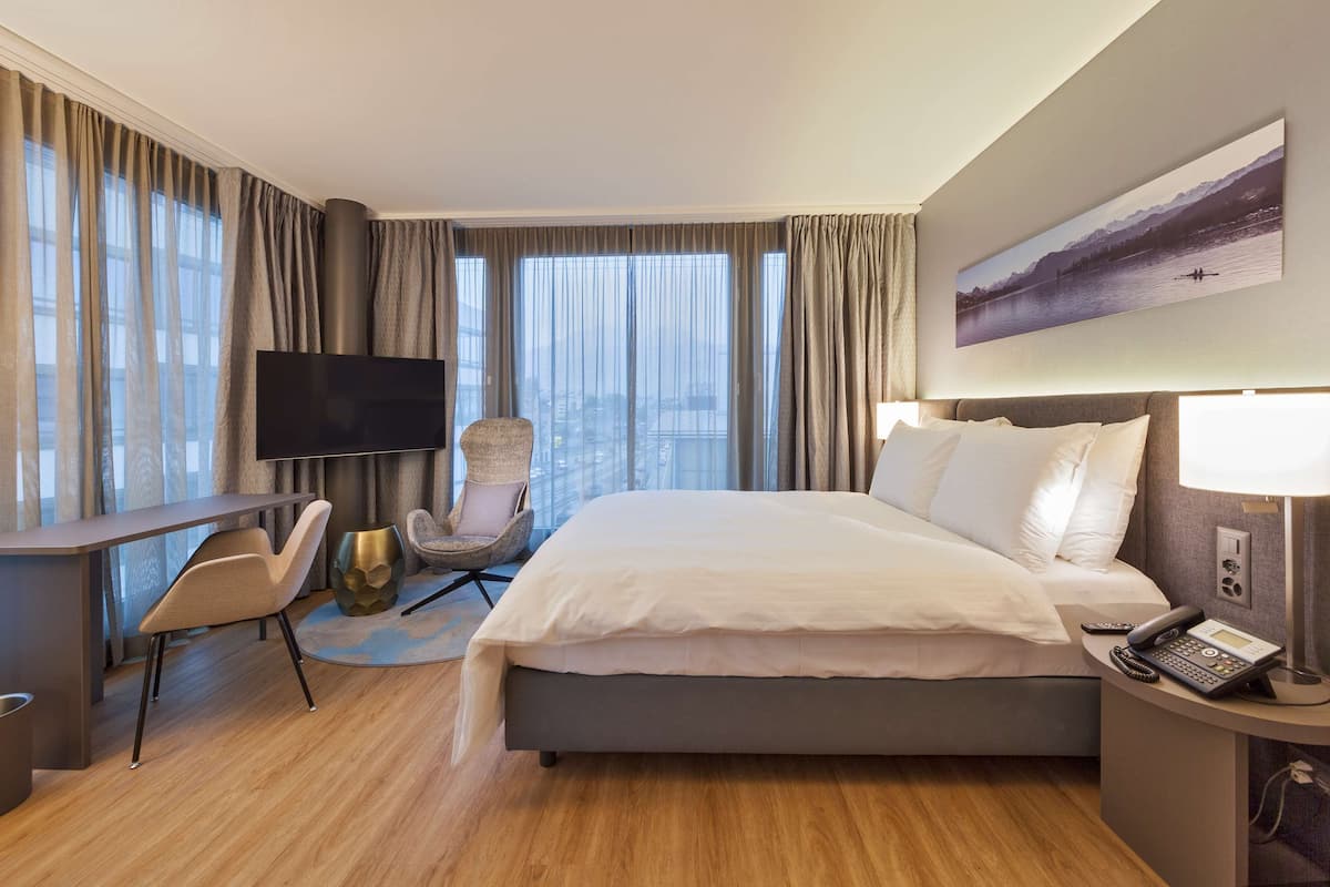 A modern hotel room with a large bed, a desk, a chair, a TV, and a painting on the wall.