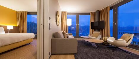 Una habitación de hotel con cama, sofá, sillón, televisor y ventanas amplias con vista a la ciudad.