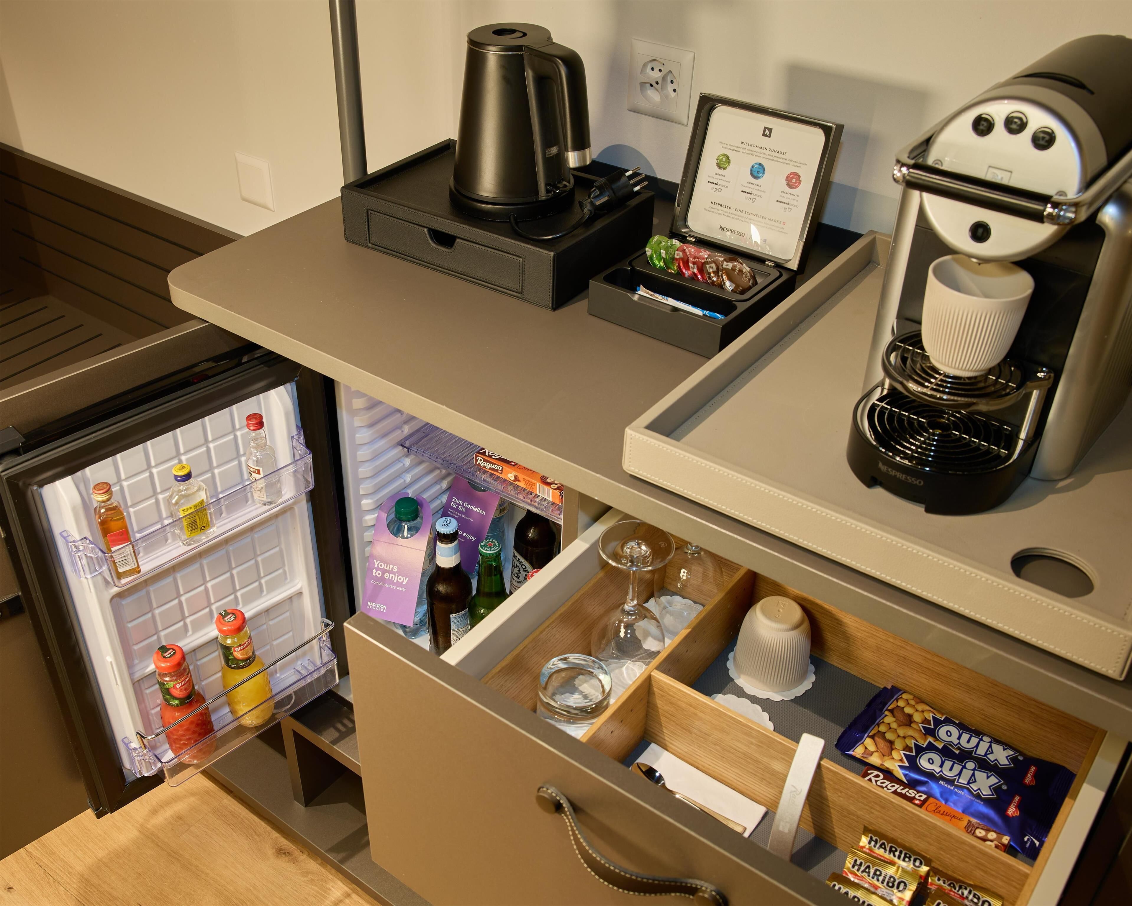 A compact kitchenette with a coffee maker, a kettle, and a mini-fridge containing beverages and condiments.