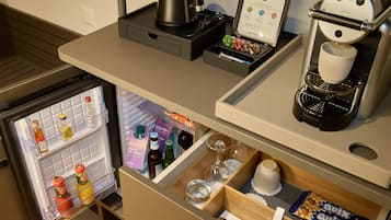 Una kitchenette compacta con cafetera, hervidor y un mini refrigerador que contiene bebidas y aderezos.