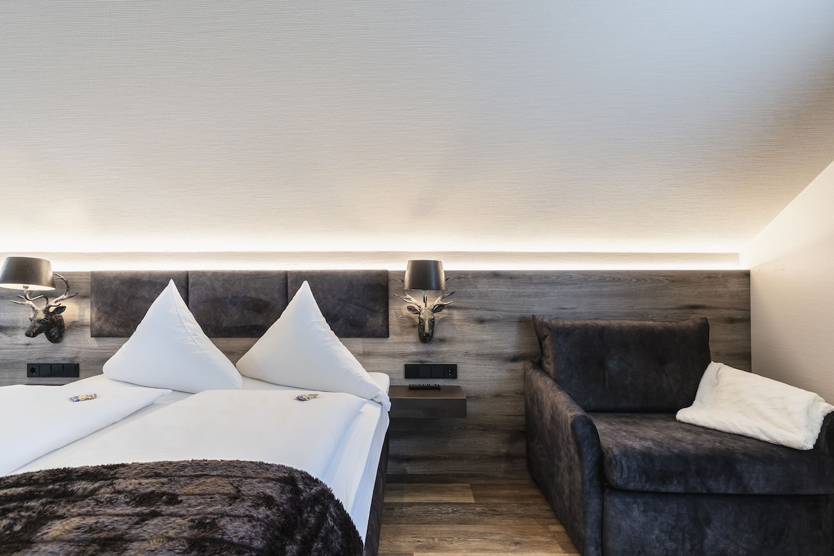 A modern bedroom with a bed, a dark armchair, and a headboard with wall-mounted lights.