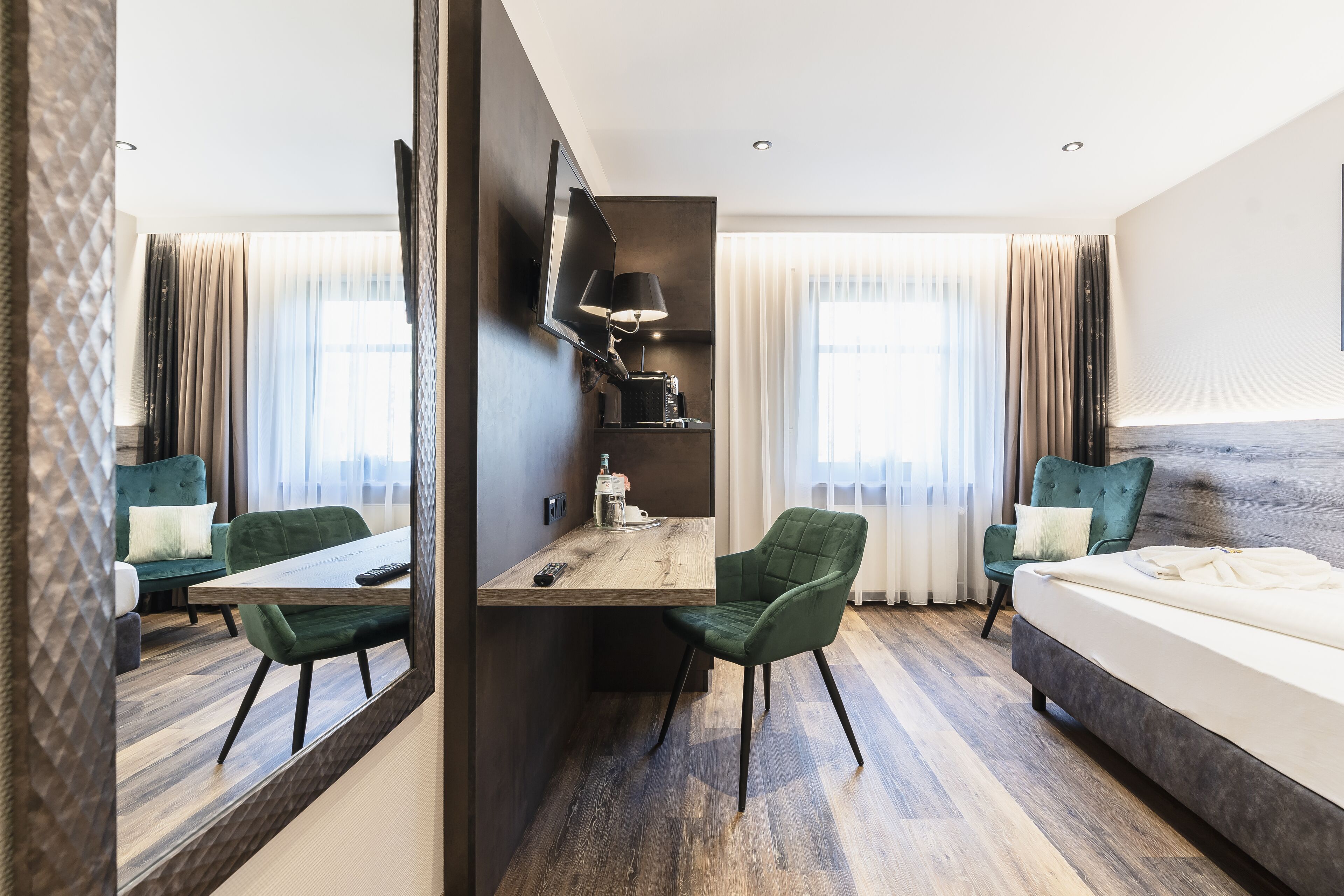 A hotel room with a wooden desk, two green chairs, a bed, a TV mounted on the wall, and a large mirror.