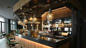 A bar area with a wooden counter, multiple hanging light fixtures, and a variety of bottles and glasses on shelves. - Hotel DEMAS Unterhaching (Unterhaching)