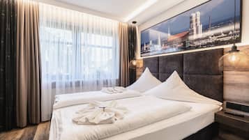 A hotel room with a large bed, two pillows, a headboard, a nightstand, a lamp, and a cityscape painting on the wall.