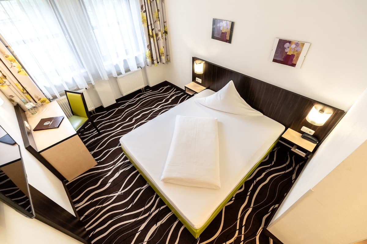 A hotel room with a zebra-patterned floor, a neatly made bed with white linens, a bedside table with a lamp, and a window with sheer curtains.