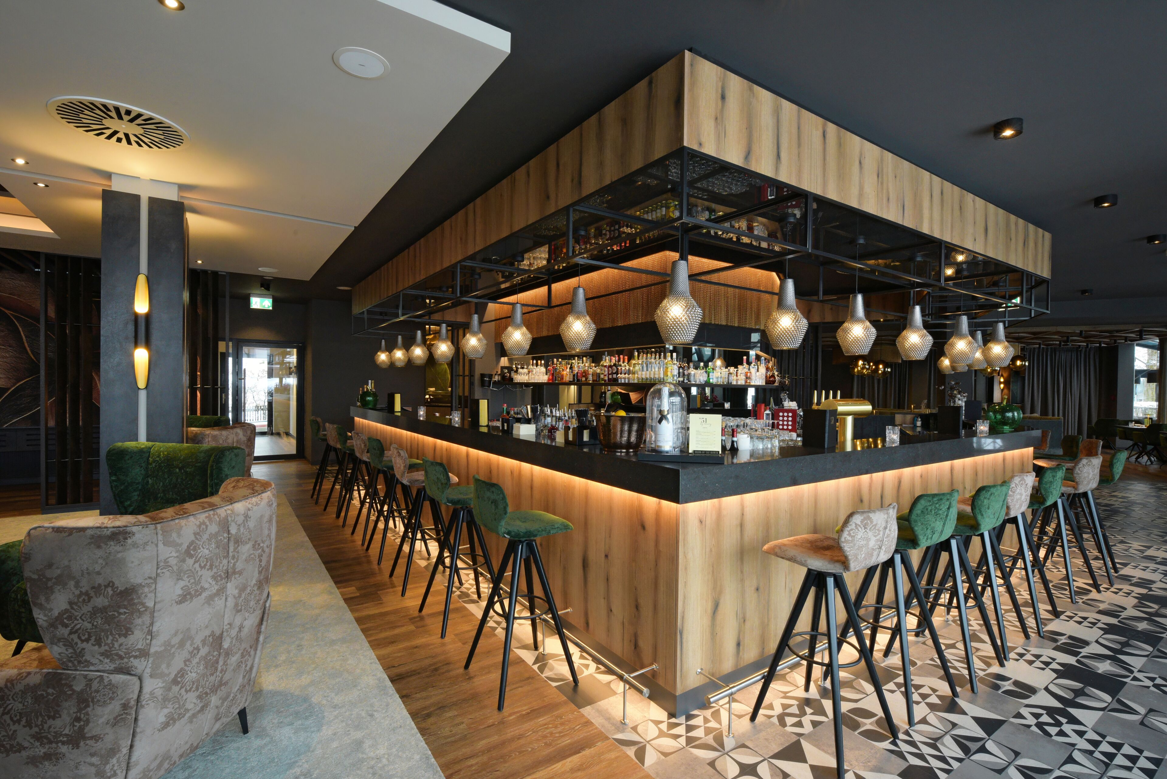 A modern bar area with a wooden counter, multiple hanging pendant lights, and green upholstered bar stools.