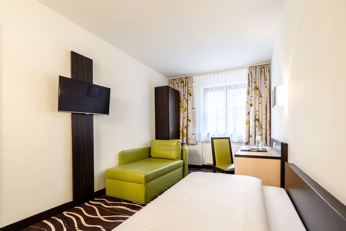 A hotel room with a bed, a green armchair, a desk with a chair, a flat-screen TV mounted on the wall, and a window with curtains.
