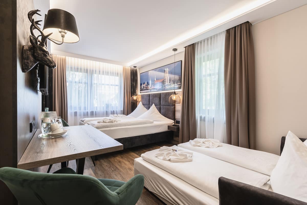 A hotel room with two beds, a dining table, a green chair, and a mounted deer head.