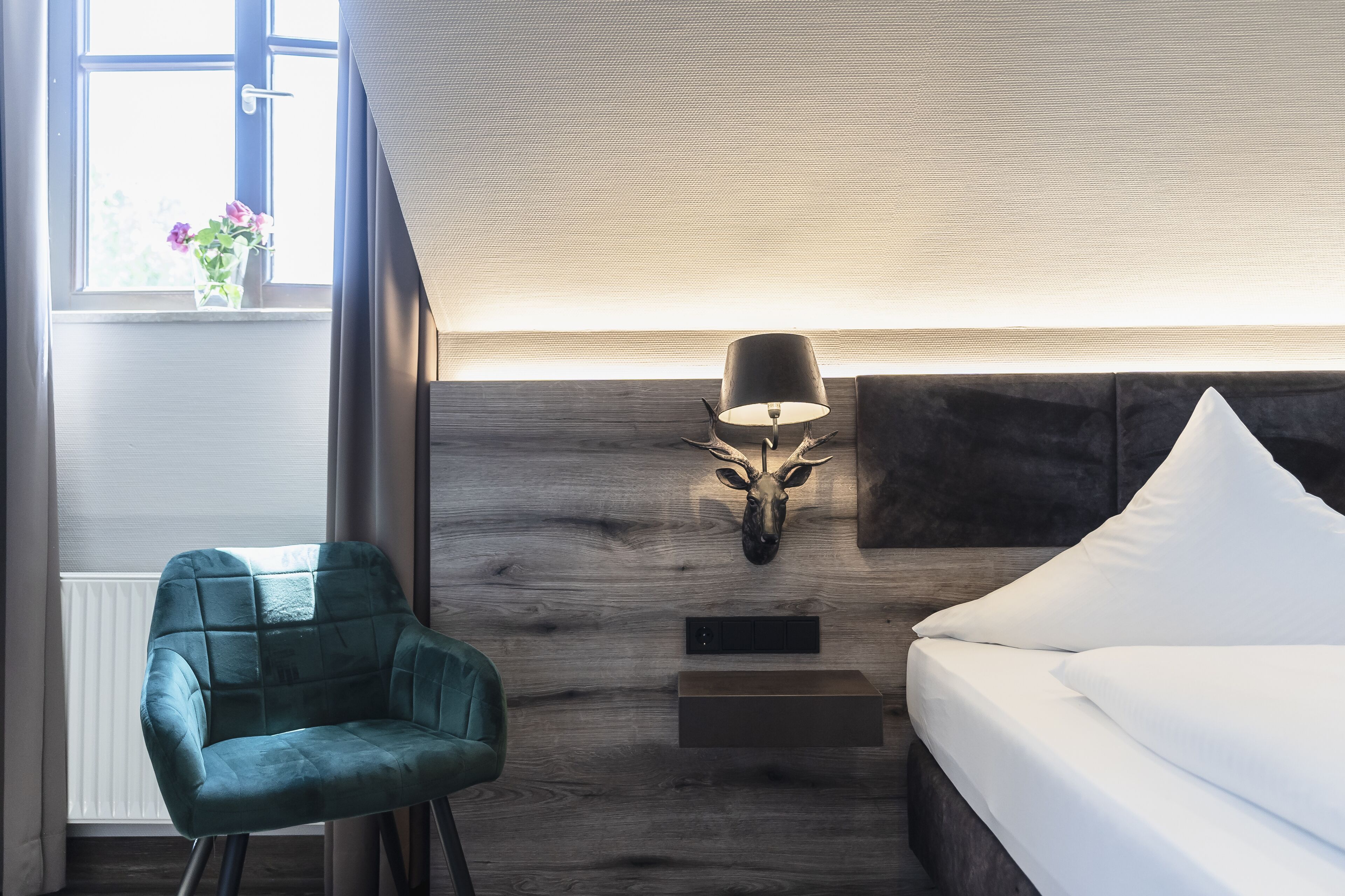 Une chambre d’hôtel moderne avec un mur en bois, une tête de cerf décorative accrochée au mur, une lampe de chevet, un fauteuil turquoise et un lit avec du linge de lit blanc.