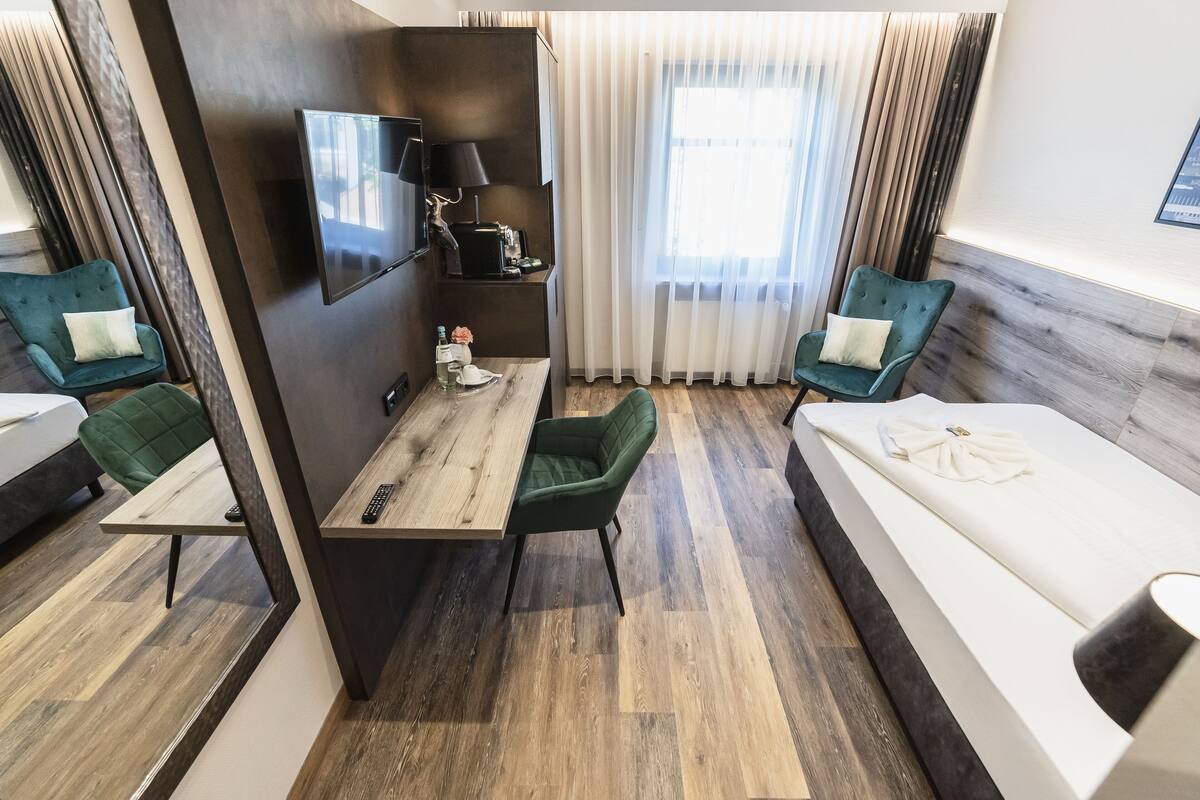 A hotel room with a wooden desk, two green chairs, a bed, a TV mounted on the wall, and a window with curtains.