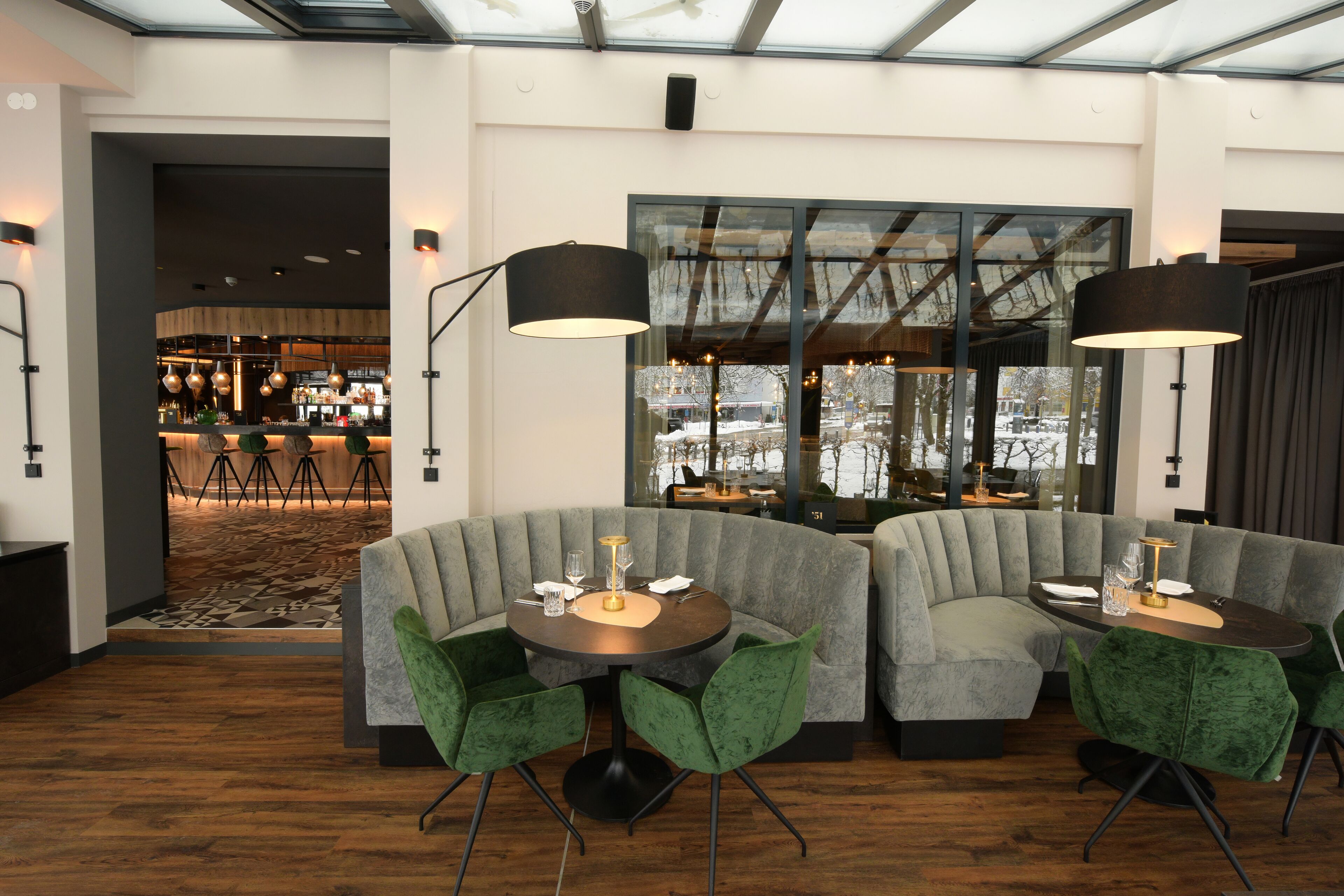 A modern dining area with a round table, green upholstered chairs, and a large window offering a view of the outdoors.