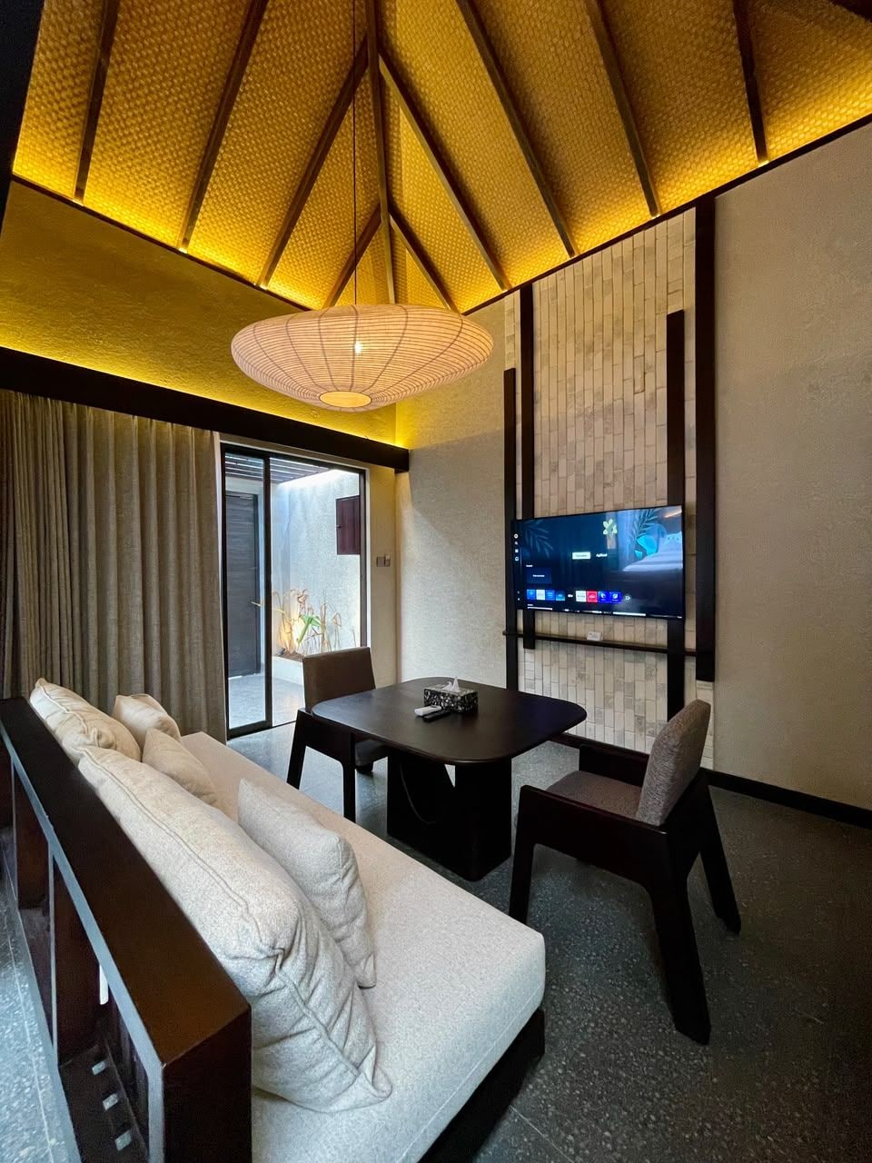 A modern living room with a sofa, dining table, chairs, a TV mounted on the wall, and a patterned ceiling.