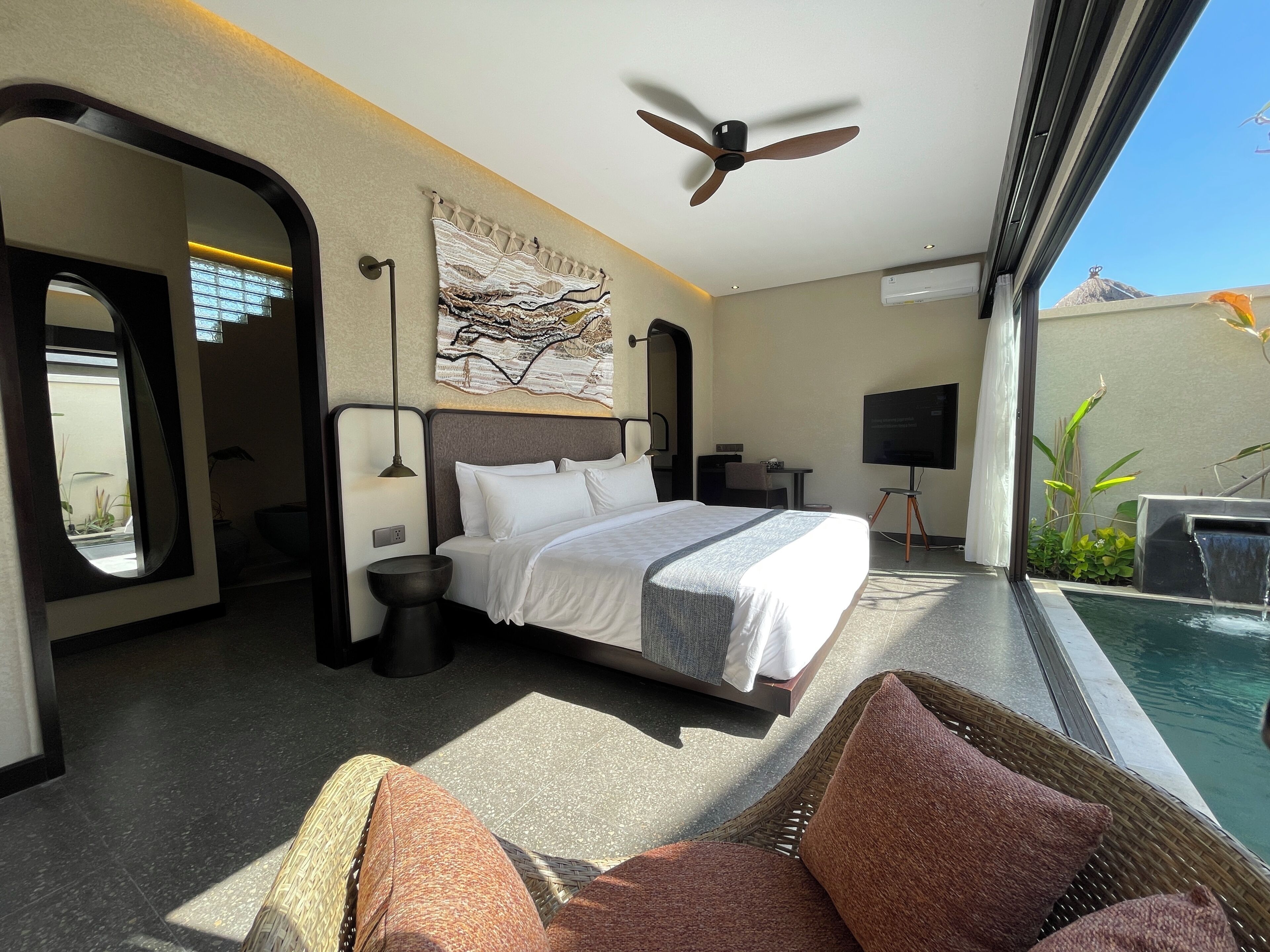 Une chambre d’hôtel avec un lit, un ventilateur de plafond, une grande fenêtre, un téléviseur fixé au mur et une vue sur une piscine.