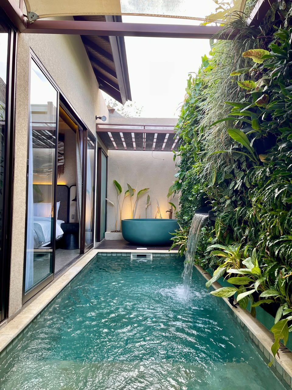 Piscina privada con caída de agua, rodeada de exuberante follaje y una puerta de cristal que da a un dormitorio.