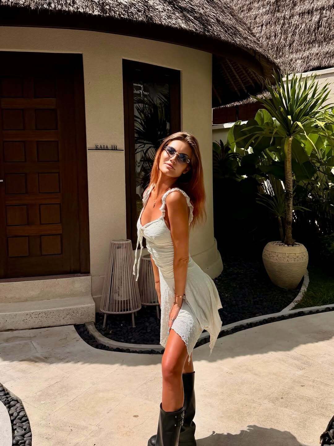 A woman in a white dress standing in front of a building with a thatched roof and a door.