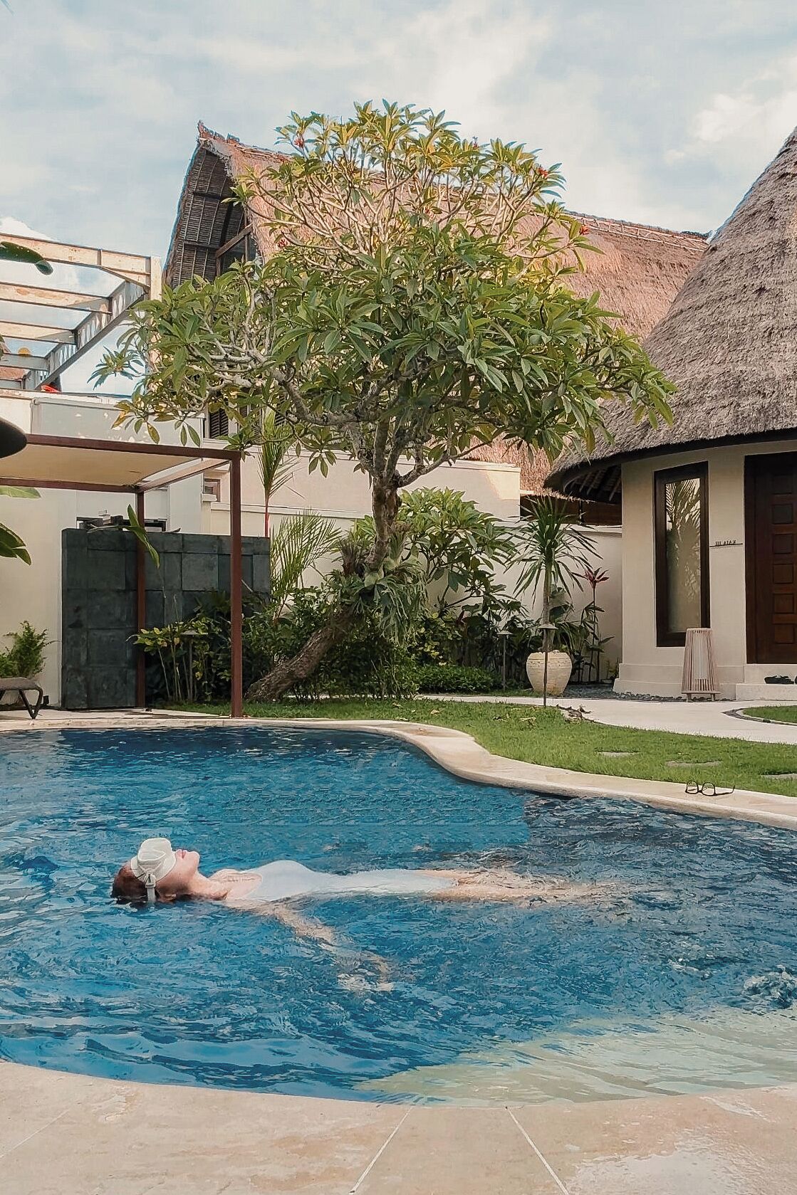 A swimming pool with a person swimming, surrounded by greenery and a building with a thatched roof.