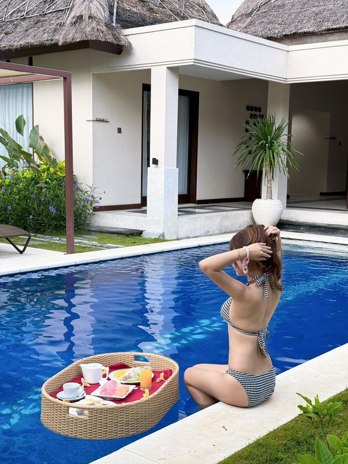 A poolside scene with a person sitting by the pool, a wicker tray with food and drinks, and a building with a thatched roof in the background.