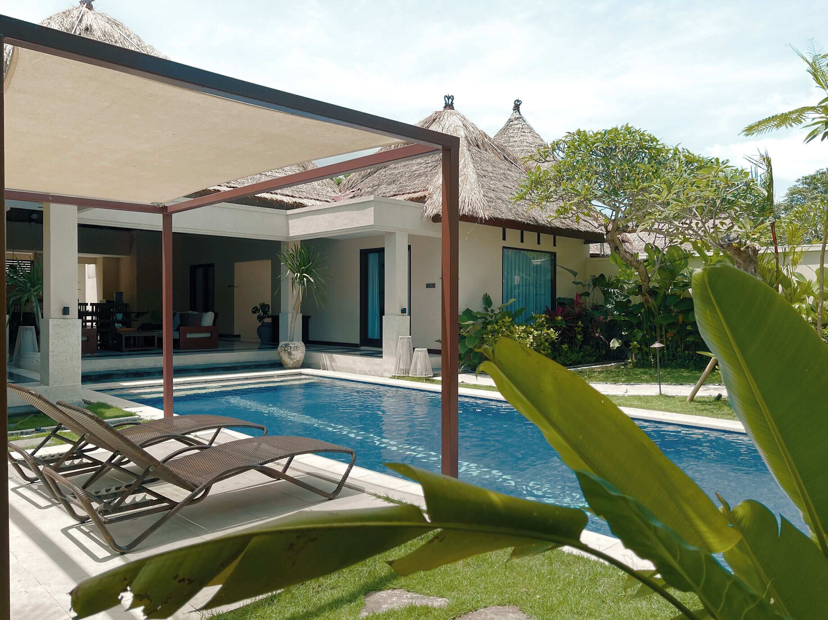 A pool area with lounge chairs, a covered seating area, and a building with thatched roofs in the background.