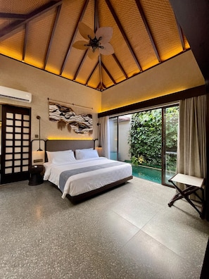 Une chambre dotée d’un grand lit, d’un ventilateur de plafond, d’une œuvre d’art accrochée au mur et d’une porte-fenêtre coulissante donnant sur un jardin.
