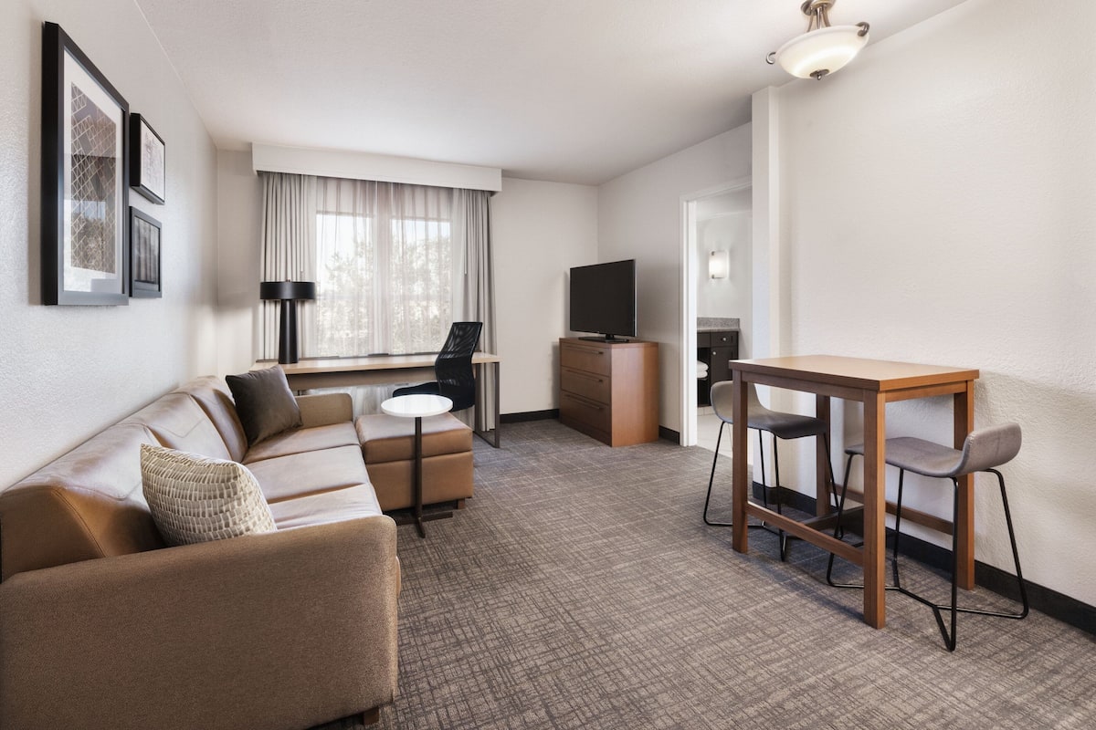 A modern hotel room with a sofa, a small dining table with chairs, a TV, and framed pictures on the wall.
