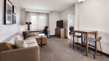 A modern hotel room with a sofa, a small dining table with chairs, a TV, and framed pictures on the wall.