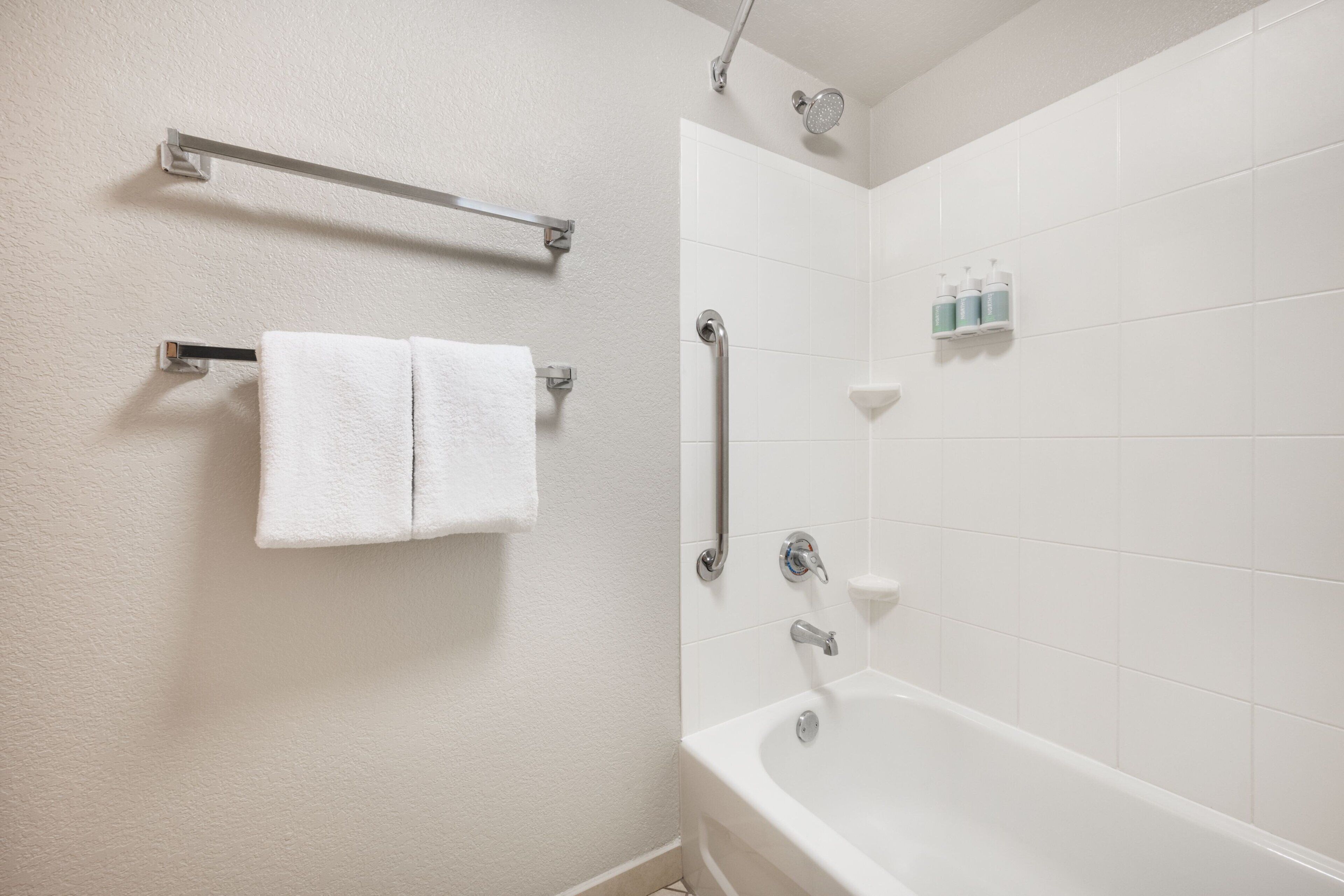 A bathroom with a white bathtub, a towel rack with two towels, a handrail, and a shelf with toiletries.