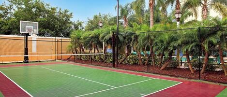 Un terrain de tennis extérieur équipé d’un panier de basket, entouré de palmiers et délimité par une clôture.