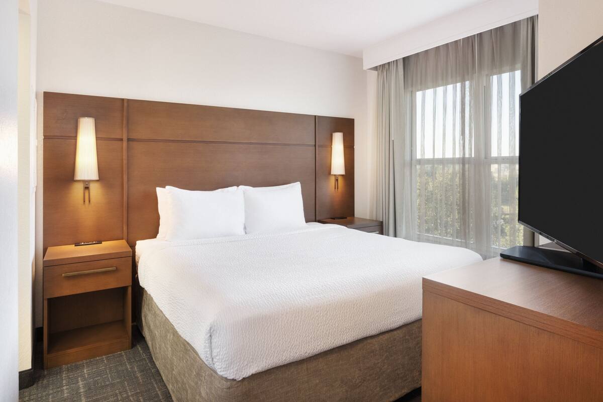 A hotel room with a large bed, two bedside tables with lamps, a flat-screen TV, and a large window with curtains.