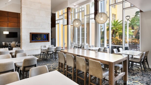 Un amplio vestíbulo de hotel con grandes ventanales, una mesa central de madera y varias zonas de estar.