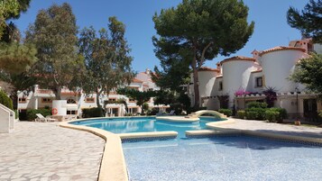 A swimming pool with a geometric tile pattern, surrounded by white buildings with red-tiled roofs, and trees.