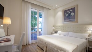 Habitación de hotel con una cama grande, un escritorio con lámpara, una silla, una ventana con cortinas y un cuadro en la pared.