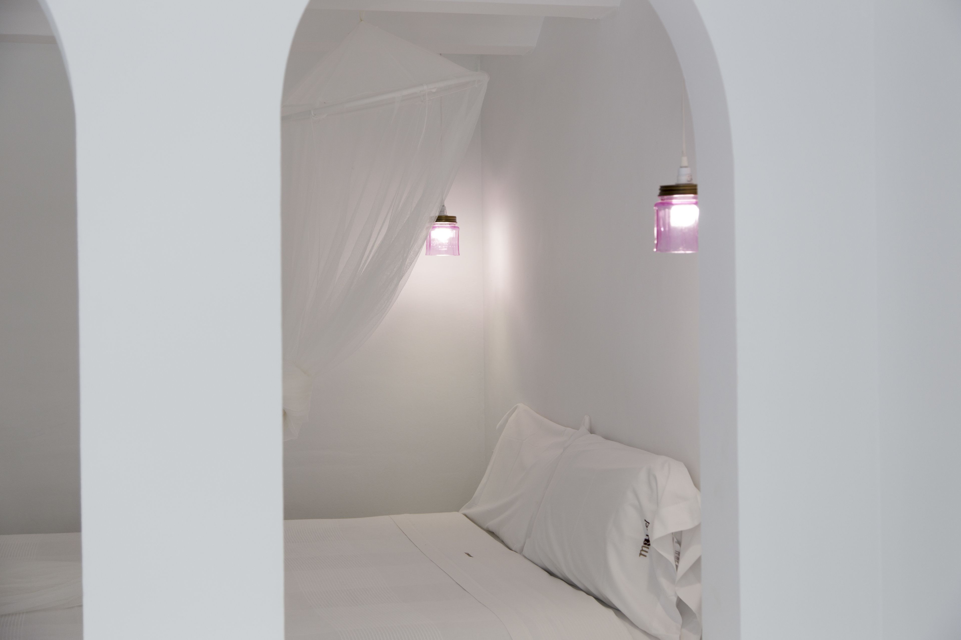 Habitació minimalista amb llit, parets blanques i dues làmpades penjades a la paret.