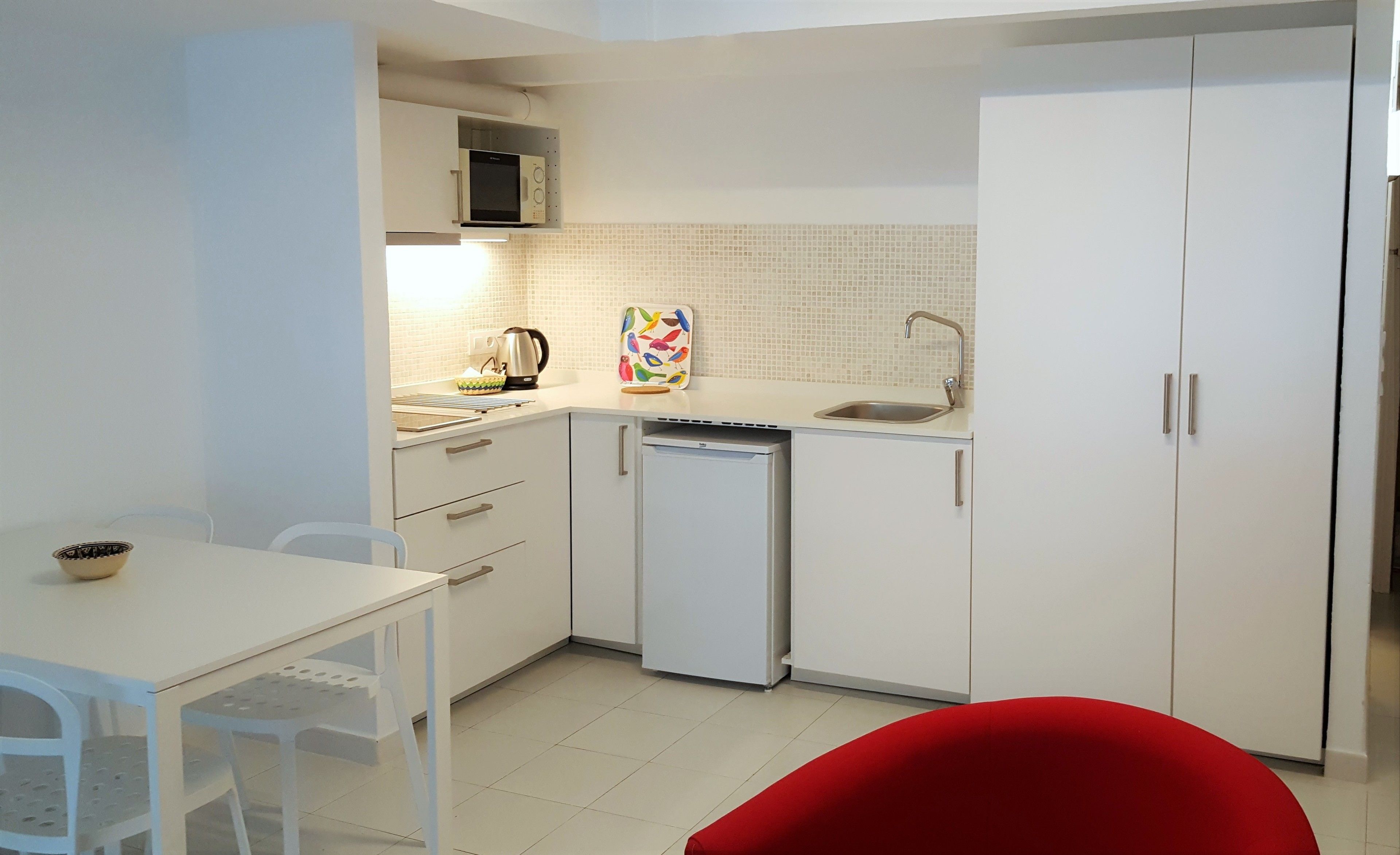 Une cuisine compacte avec des meubles blancs, un four à micro-ondes, un évier et un lave-vaisselle. On y trouve une table à manger avec un saladier et une chaise.
