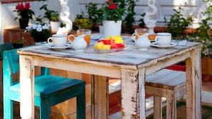 Un área de comedor al aire libre de estilo rústico, con una mesa de madera preparada para el desayuno, rodeada de plantas en macetas y un edificio blanco con ventanas amplias.