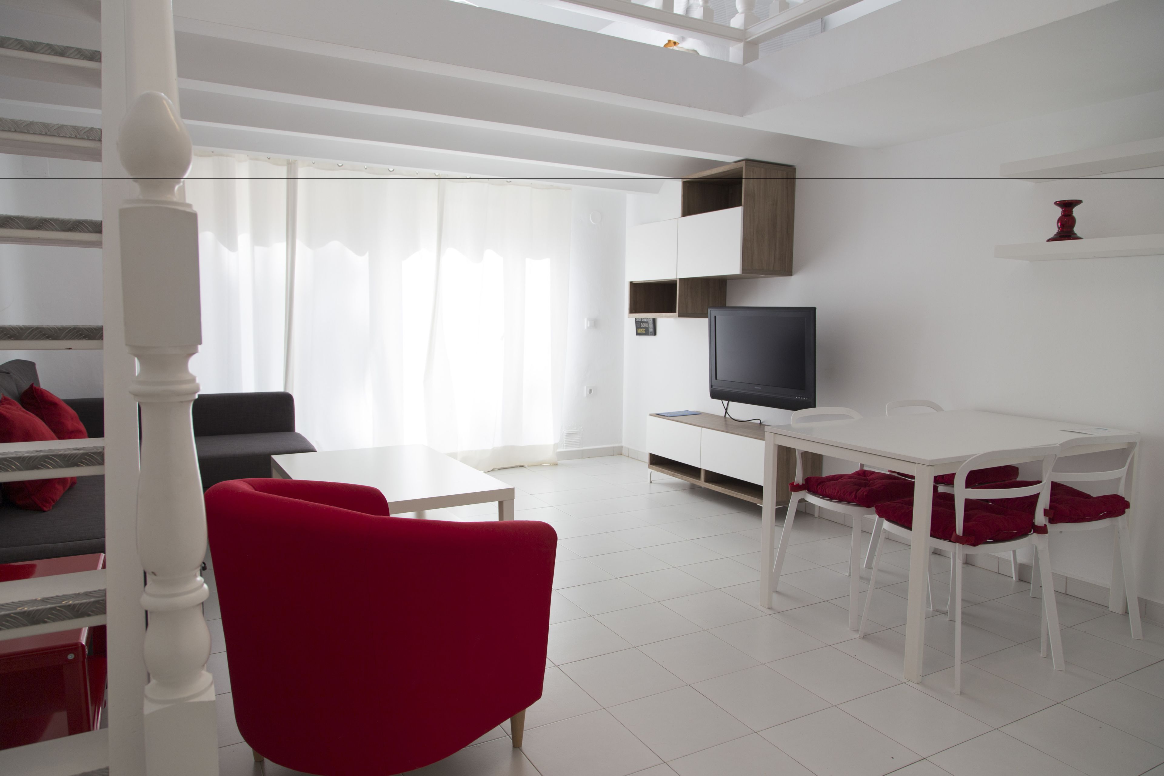 A modern living room with a white staircase, a flat-screen TV, a dining table with chairs, and a sofa with red cushions.