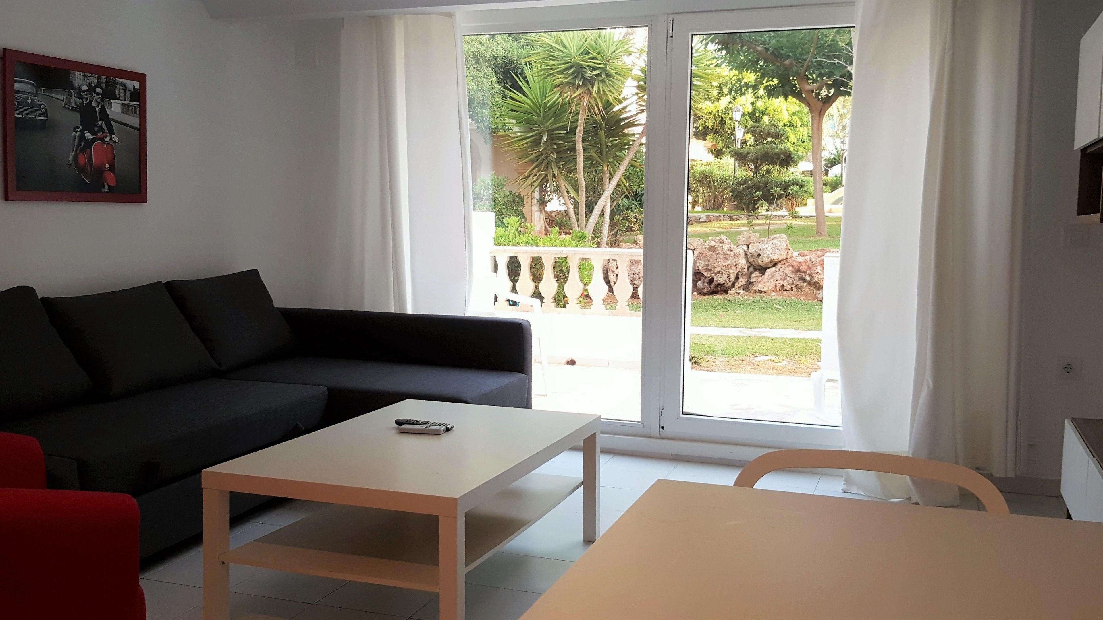 Un salón con un sofá negro, una mesa blanca y una silla roja. Hay un ventanal corredero que da a un jardín con una palmera y una valla.