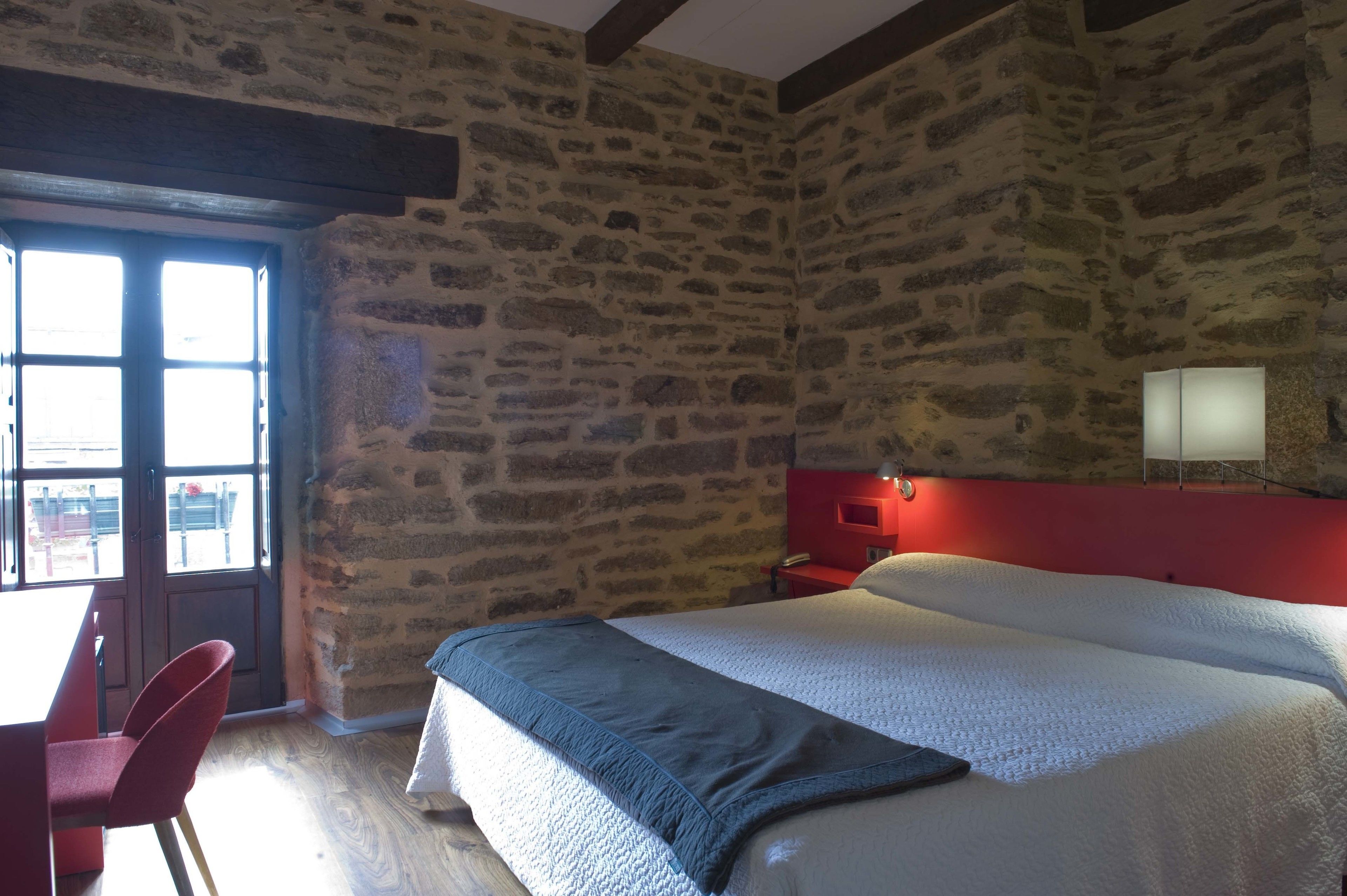 Una habitación con pared de piedra, una cama con manta azul, cabecero rojo, una mesita de noche y una ventana con vistas al exterior.