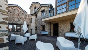 Un edificio de piedra con una terraza de madera, mobiliario exterior blanco y un balcón con barandilla.