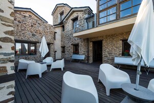 Un edificio de piedra con una terraza de madera, mobiliario exterior blanco y un balcón con barandilla.