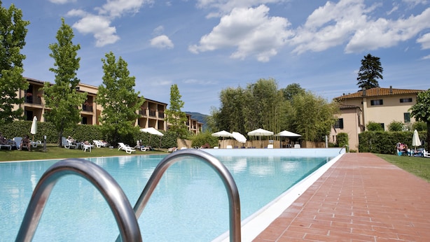 A swimming pool with a tiled deck, surrounded by lounge chairs and umbrellas, with buildings in the background. - Hotel Villa Cappugi (Pistoia)