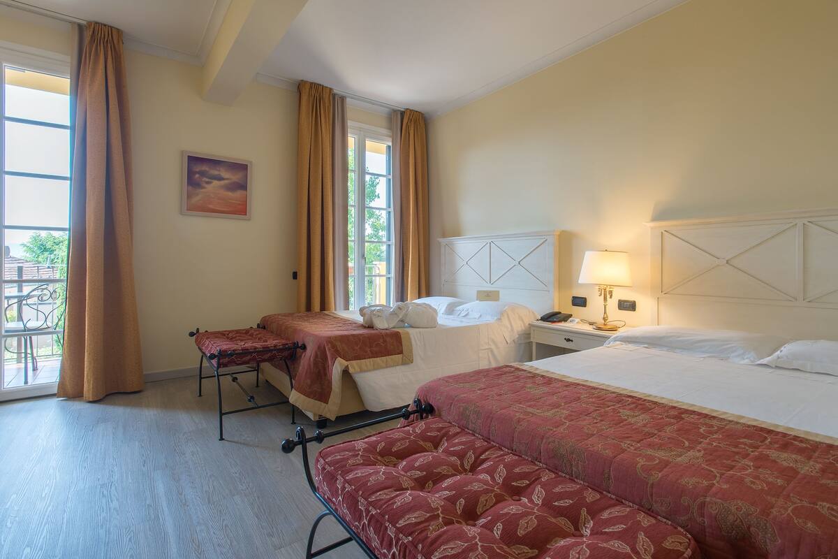 A hotel room with two beds, a bench, a window with curtains, a bedside table with a lamp, and a painting on the wall.