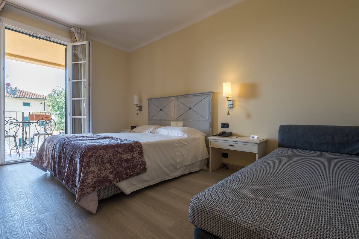 A hotel room with a bed, a desk, and a chair. There is a balcony with a table and chairs outside.