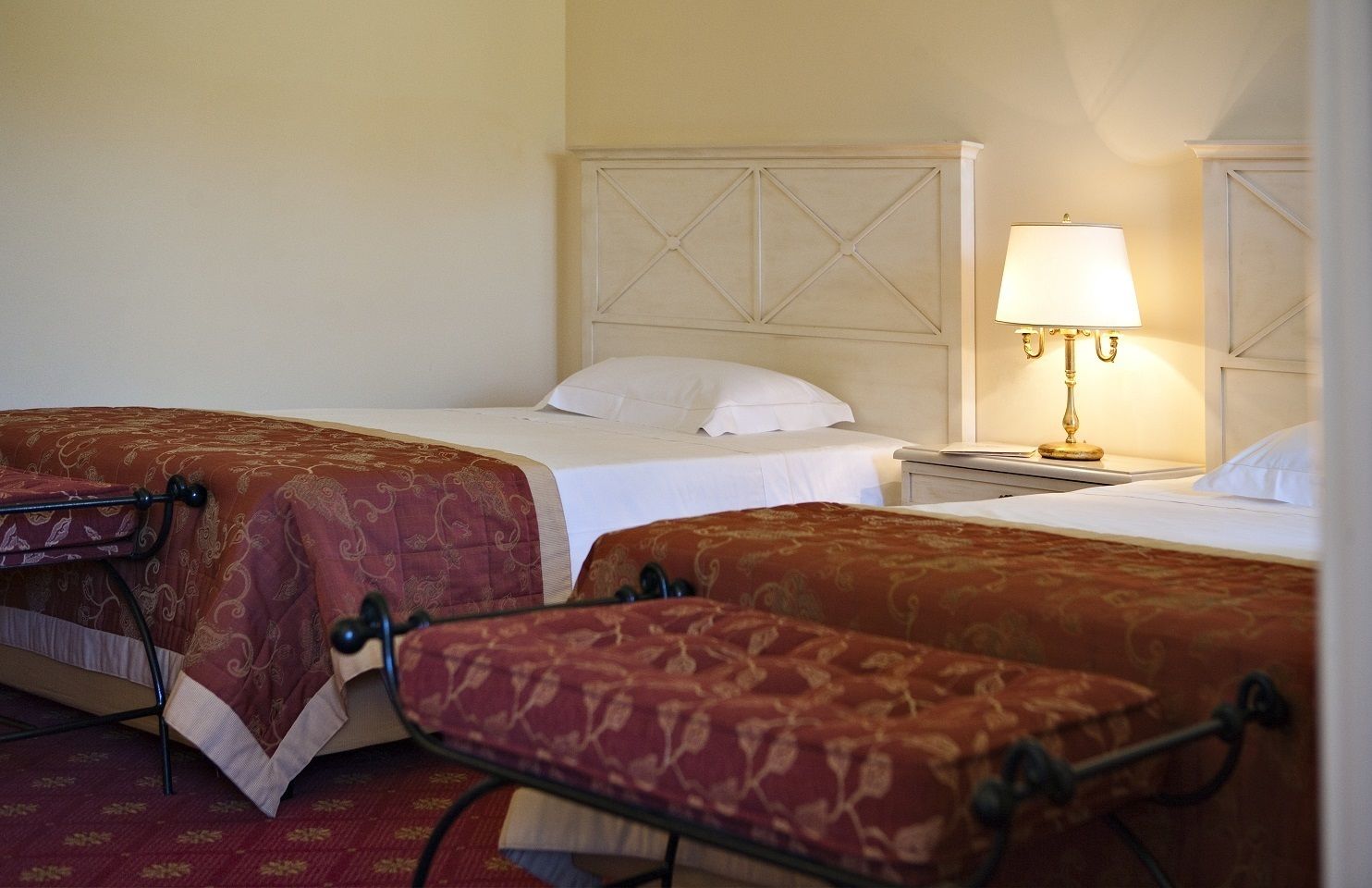 Une chambre d’hôtel avec deux lits, chacun recouvert d’une couvre-lit à motifs rouges et agrémenté d’oreillers blancs. Une table de chevet avec une lampe se trouve entre les deux lits.