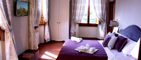 A neatly arranged bedroom with a bed, a bedside table, a window with curtains, a wall-mounted TV, and a small stool.