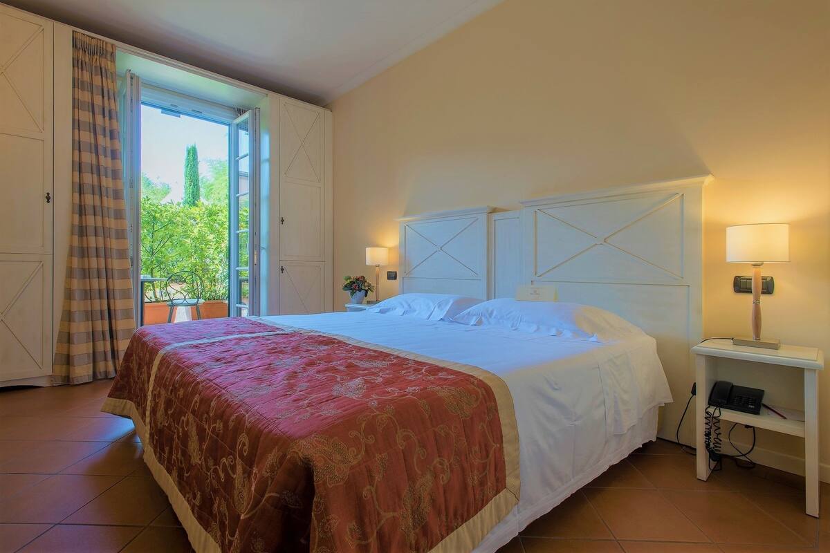 A hotel room with a large bed, a nightstand with a lamp, and a view of greenery through the open door.