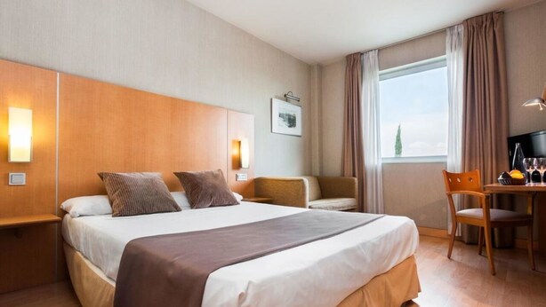 Een hotelkamer met een groot bed, een bureau met stoel, een raam met gordijnen en een aan de muur bevestigde televisie.