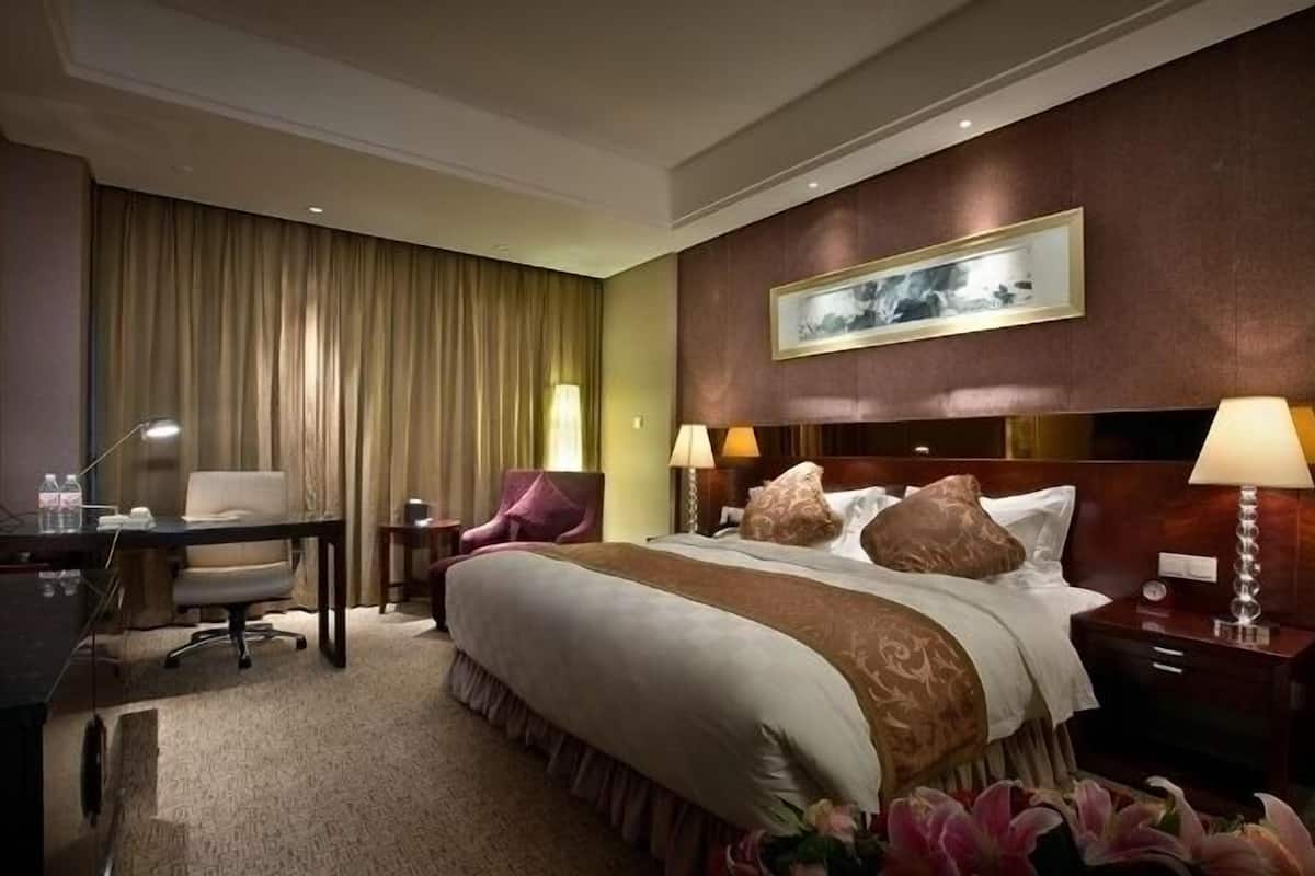Ein Hotelzimmer mit einem großen Bett, zwei Nachttischen mit Lampen, einem Schreibtisch mit Stuhl, einem großen Fenster mit Vorhängen und einem gerahmten Bild an der Wand.