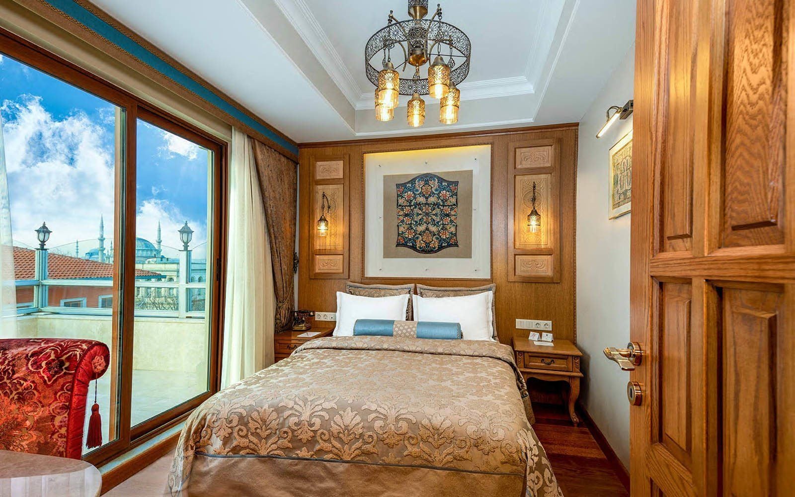 A hotel room with a large bed, wooden paneling, a chandelier, and a view of a cityscape.
