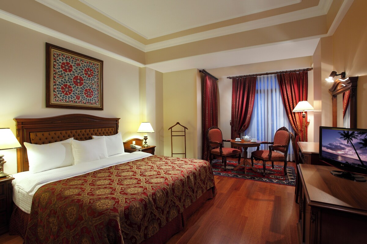 A hotel room with a large bed, a desk with a television, a chair, a table with a lamp, and a window with curtains.